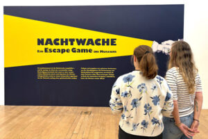 Nachtwache Escape Game im Landesmuseum Bonn, zwei Frauen schauen das große Logo der Nachwache an